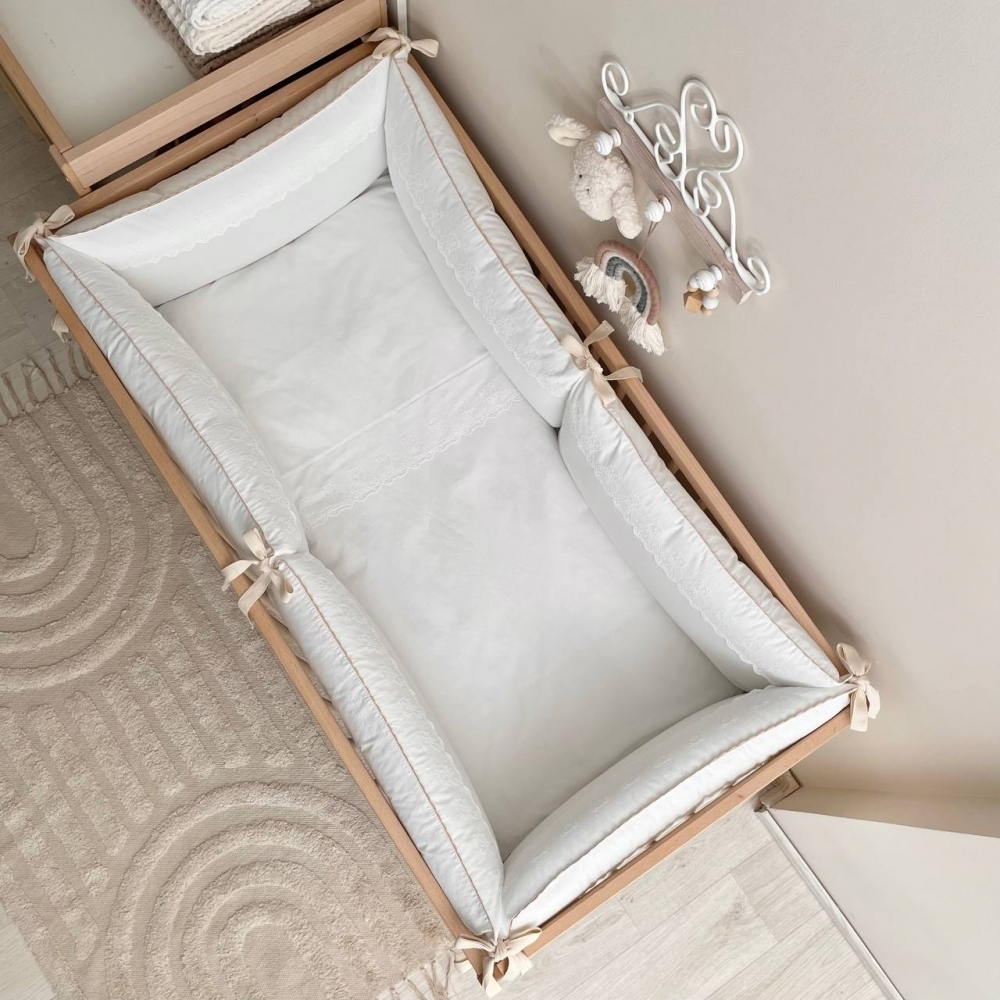 Ansicht von oben auf das Babybett mit dem Bettwäsche-Set, das die Decke, das Kissen und die Bumper in Milchweiß zeigt, verziert mit einem süßen Teddy-Motiv für ein gemütliches, stilvolles Ambiente